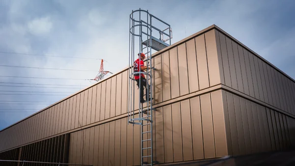 Arbeiter steigt über EASiTec Steigtechnik Steigleitern an Gebäudefassade mit Rückenschutz sicher auf das Flachdach hinauf.
